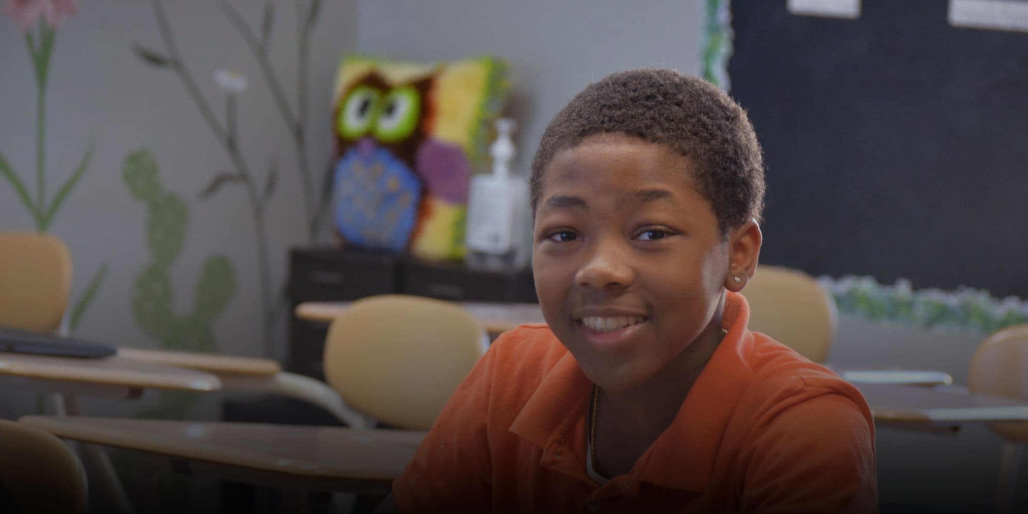 Smiling student in an orange polo shirt at a classroom desk.